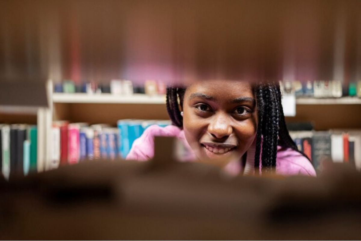 Uma menina na biblioteca é vista pela fresta entre as estantes. Ela procura livros para estudar para a Prova ETEC 2025