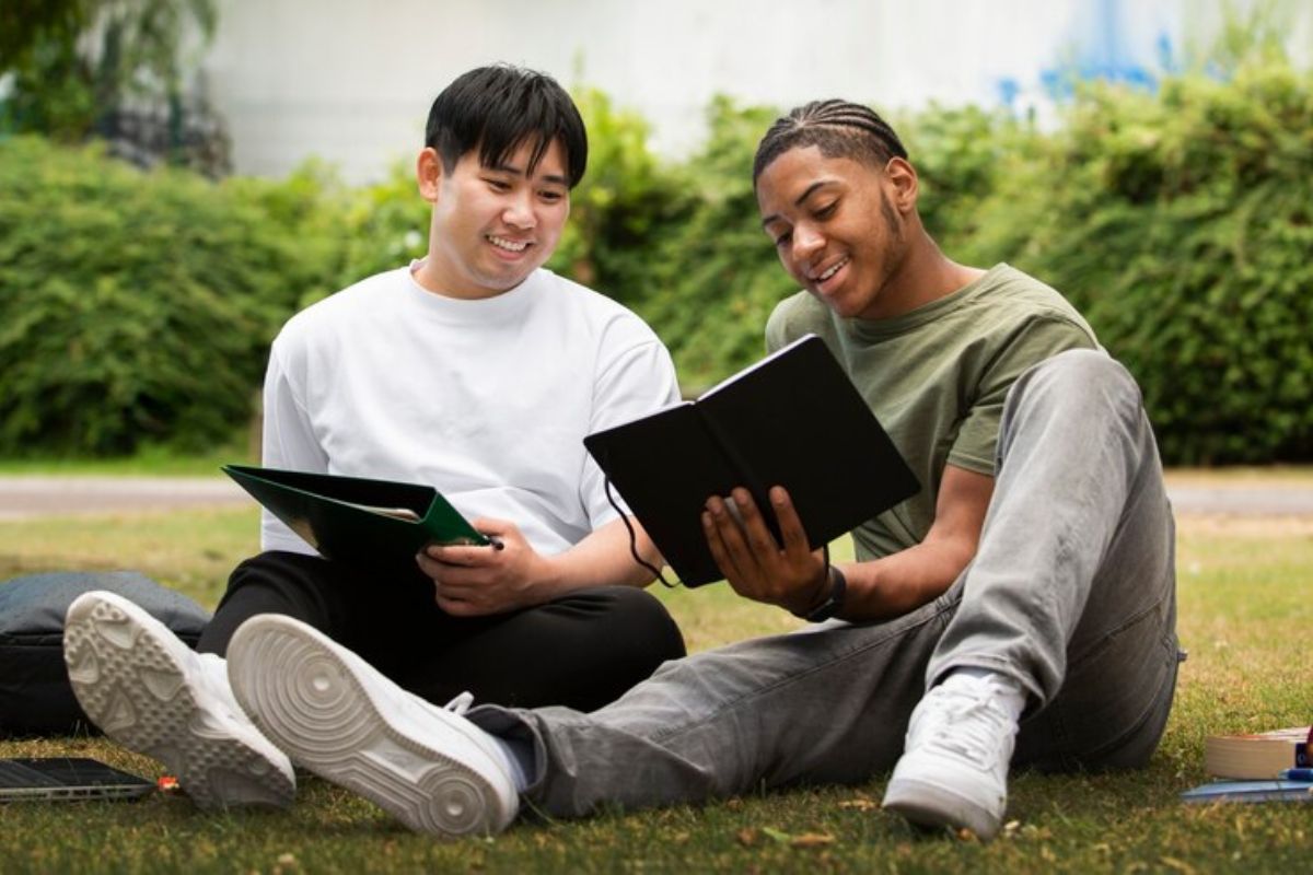 Dois alunos sentados no jardim da faculdade conversam com livros abertos nas mãos.