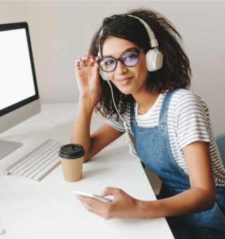 A estudante sentada em frente ao seu computador, olha para a foto. Ela usa um headphone enquanto pesquisa informações sobre o Prouni e SISU 2025