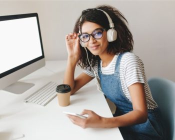 A estudante sentada em frente ao seu computador, olha para a foto. Ela usa um headphone enquanto pesquisa informações sobre o Prouni e SISU 2025