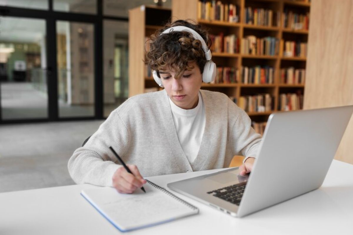Um estudante na biblioteca faz anotações em seu caderno. Ele usa um headphone e olha para o notebook que está em sua frente