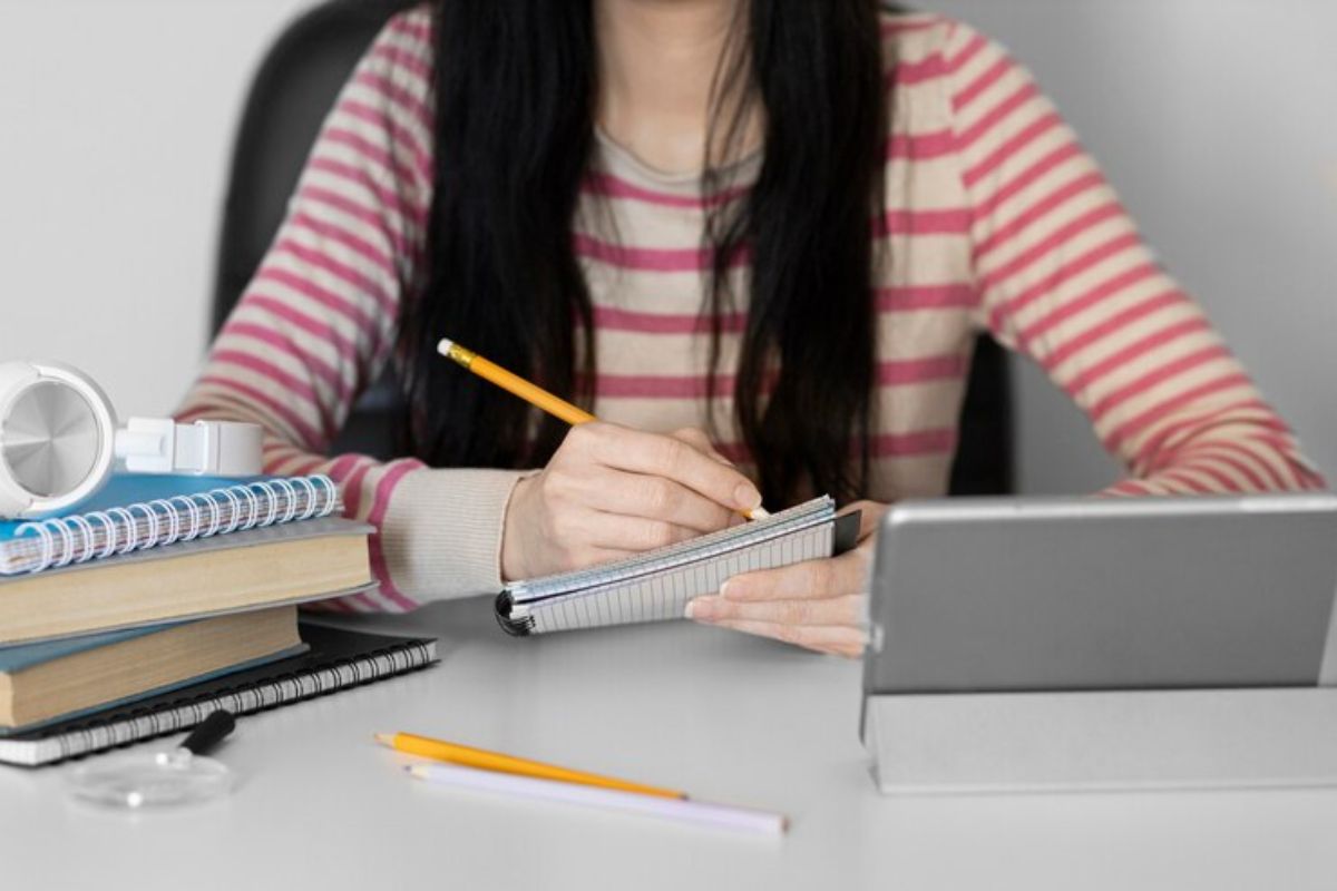 Uma menina estuda em casa. Na mesa, além do notebook, há diferentes materiais para estudo