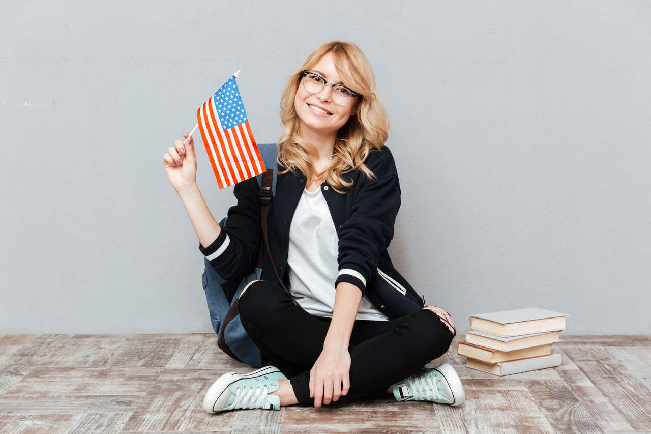 Jovem estudante de intercâmbio sentada no chão ao lado de livros posa sorridente segurando bandeira dos Estados Unidos