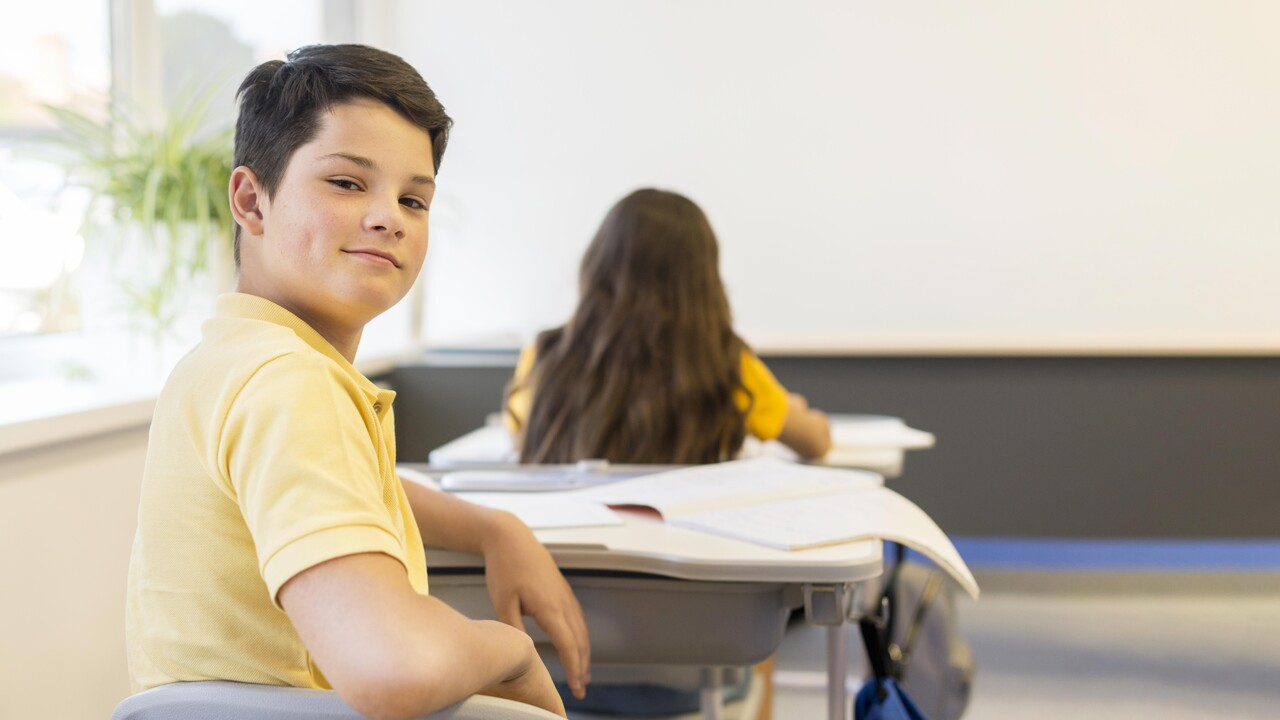 Menino posa pra foto olhando para trás sentado em sua mesa na sala de aula