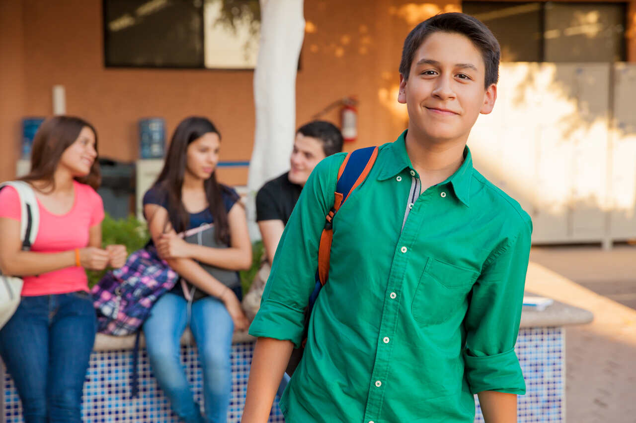 Adolescente posa sorridente no campus da escola com colegas ao fundo