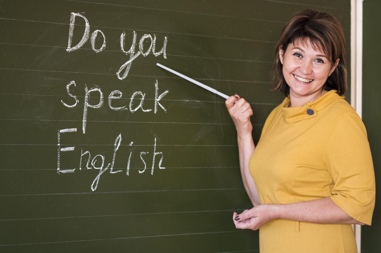 Professora de inglês posa sorridente ao lado da lousa onde está escrito: Do you speak english?