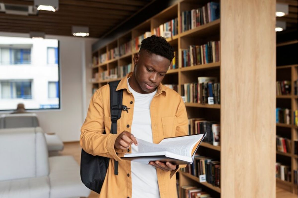 Um jovem está na biblioteca lendo um livro de pé no corredor. Ele estuda para conseguir uma vaga no Curso de Eventos ETEC 2025