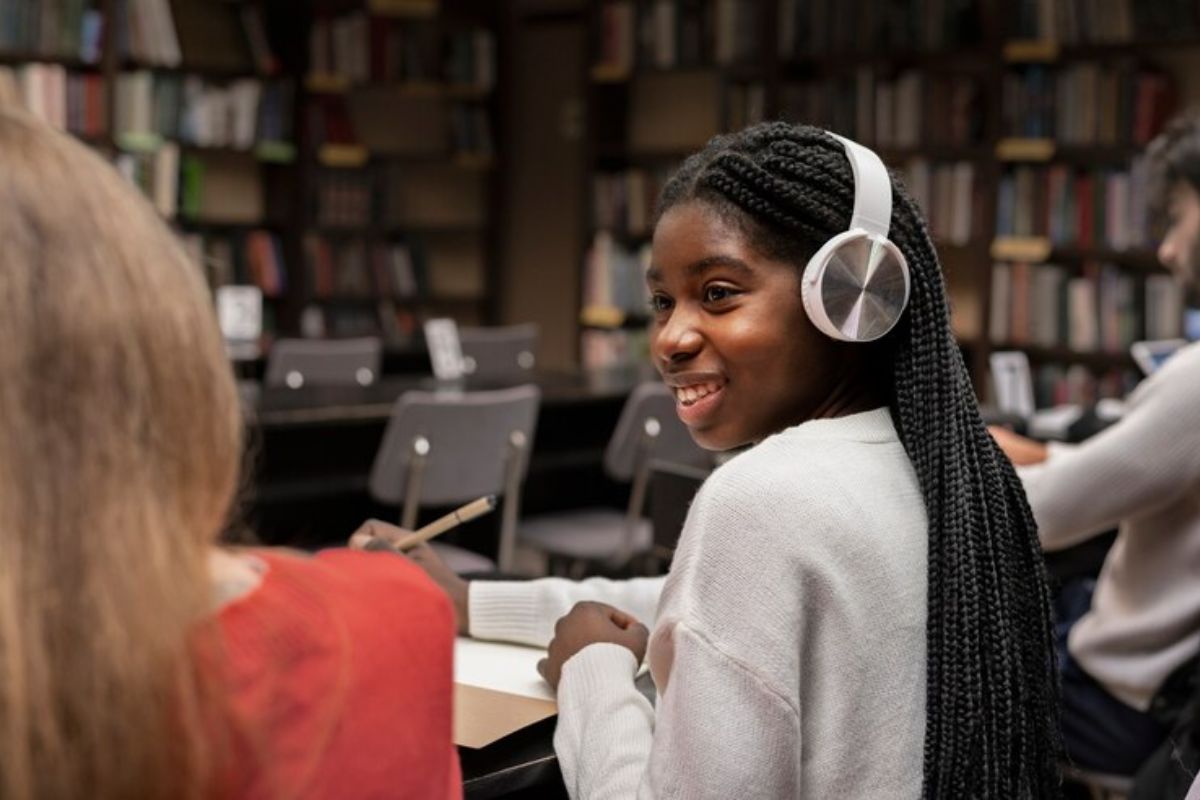 Uma menina na biblioteca olha para amiga ao lado. Ela usa um headphone enquanto mexe em seu notebook na mesa