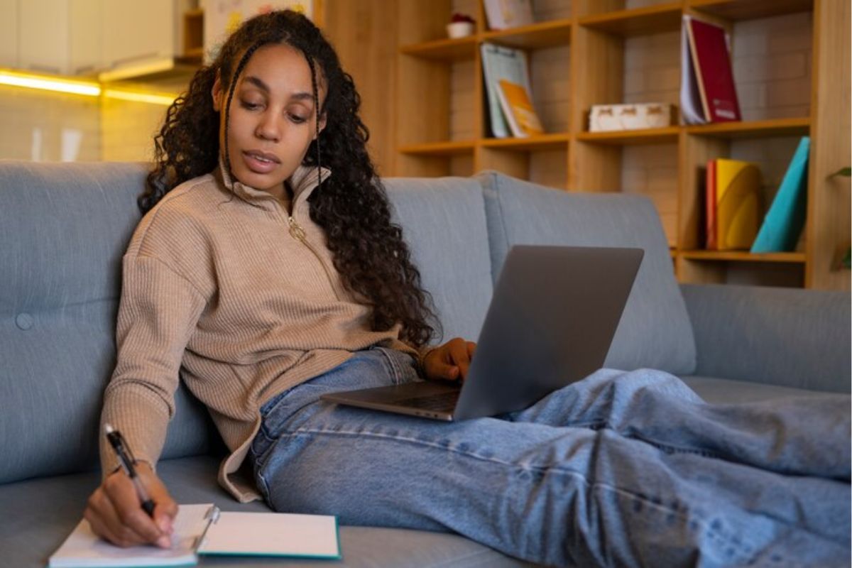 Uma estudante no sofá faz anotações no seu caderno. No notebook que está em seu colo, ela vê quando abrem as Inscrições da ETEC 2025