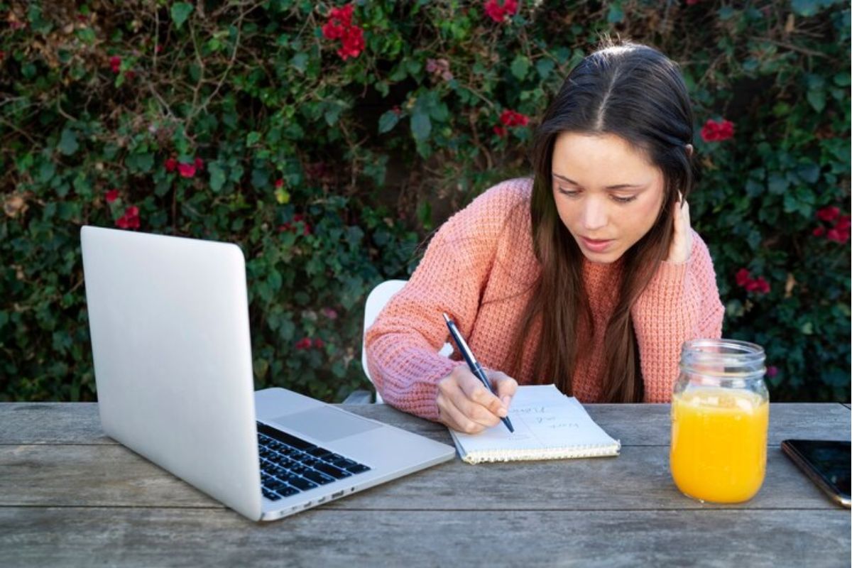 Uma menina sentada no quintal da sua casa em uma mesa. Ela faz anotação em seu caderno enquanto olha para o notebook. Na mesa também há uma jarra com suco de laranja