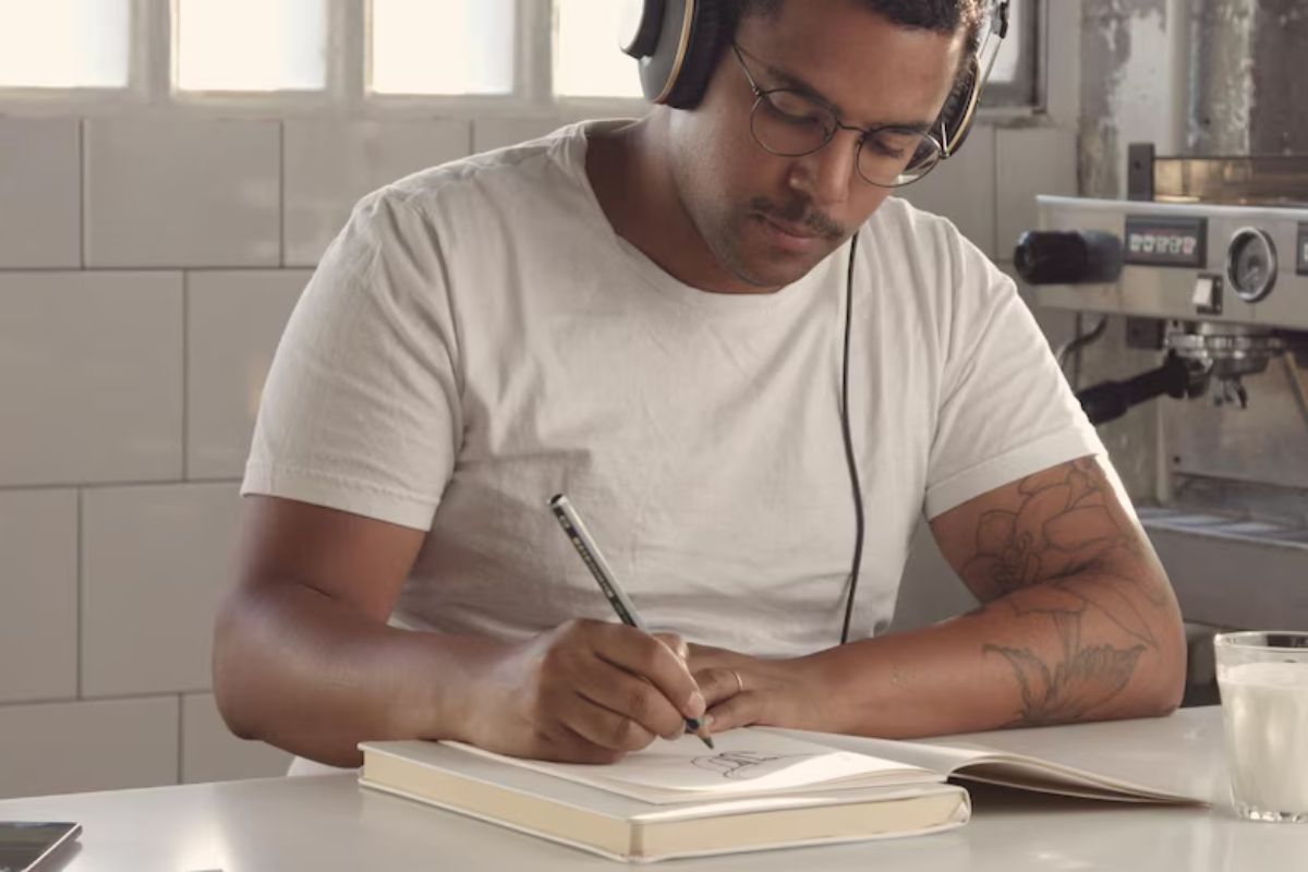 Um estudante na cozinha de sua casa escreve no caderno. Ele escuta algo no seu headphone
