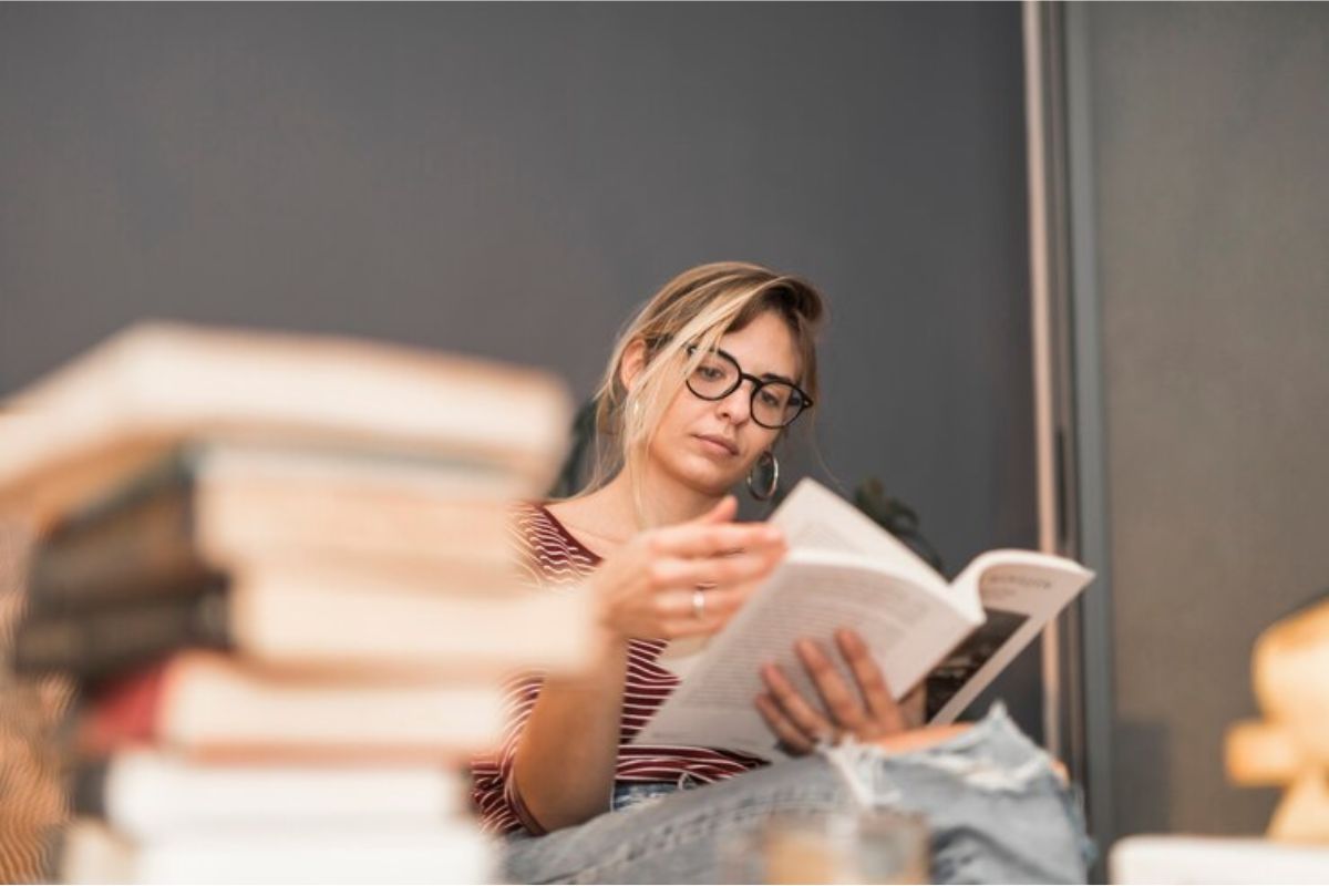 Uma estudante sentada lendo um livro. Na mesa é possível ver uma pilha de livros