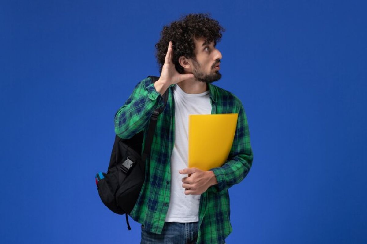 Um estudante segura seu material no colo e faz uma pose para a foto com a mão no ouvido, como se quisesse ouvir algo distante