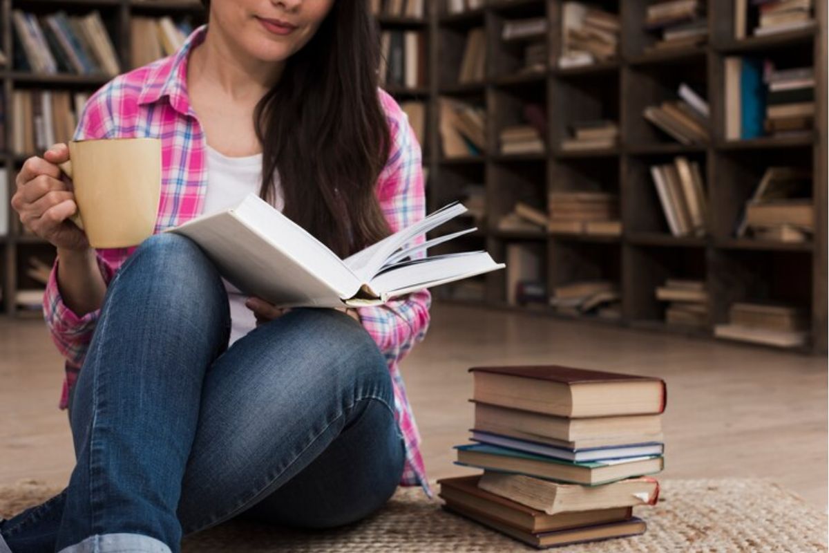 Uma estudante sentada com um livro aberto na mão. Ela está no chão e estuda segurando uma xícara 