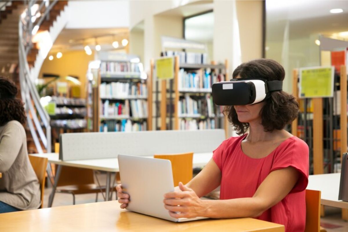 Uma estudante com óculos de Realidade Virtual. Ela está na biblioteca, sentada na mesa enquanto estuda com seu notebook