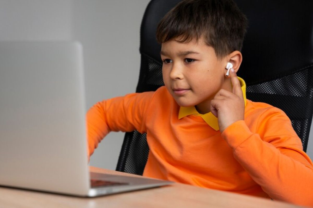 Um estudante sentado em frente a um notebook na mesa. Ele assiste um curso de robótica para crianças e escuta usando um air pod