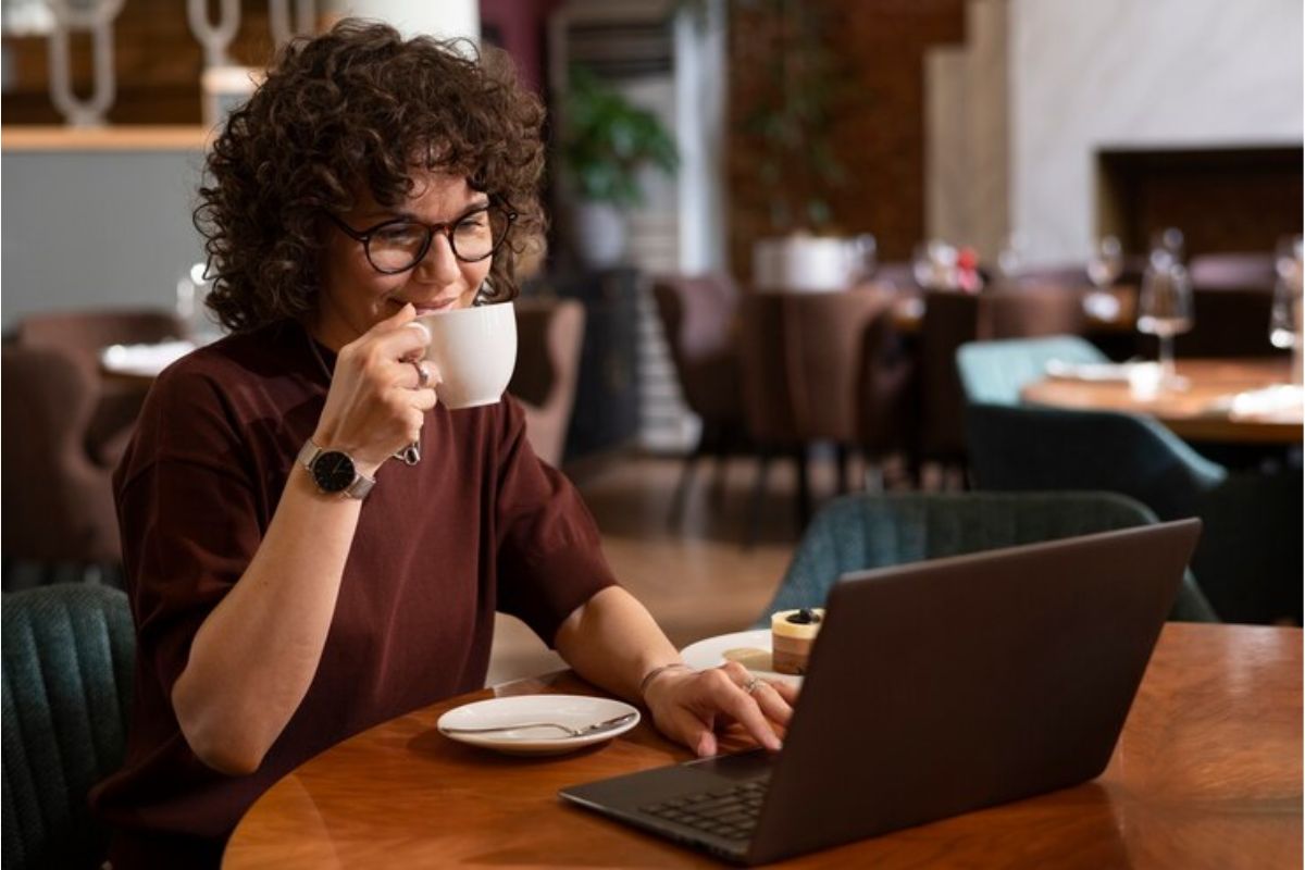 Uma estudante usa seu notebook enquanto toma café em sua xícara. Ela pesquisa pelos cursos gratuitos FGV