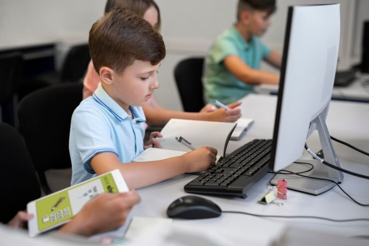 Uma criança dentro da sala de informática na frente de um computador. É possível ver outros dois estudantes na sala 