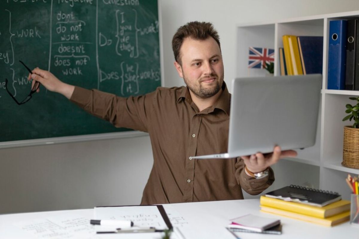 Um professor de inglês em casa. Ele segura o notebook com uma mão enquanto aponta algo na lousa atrás dele 