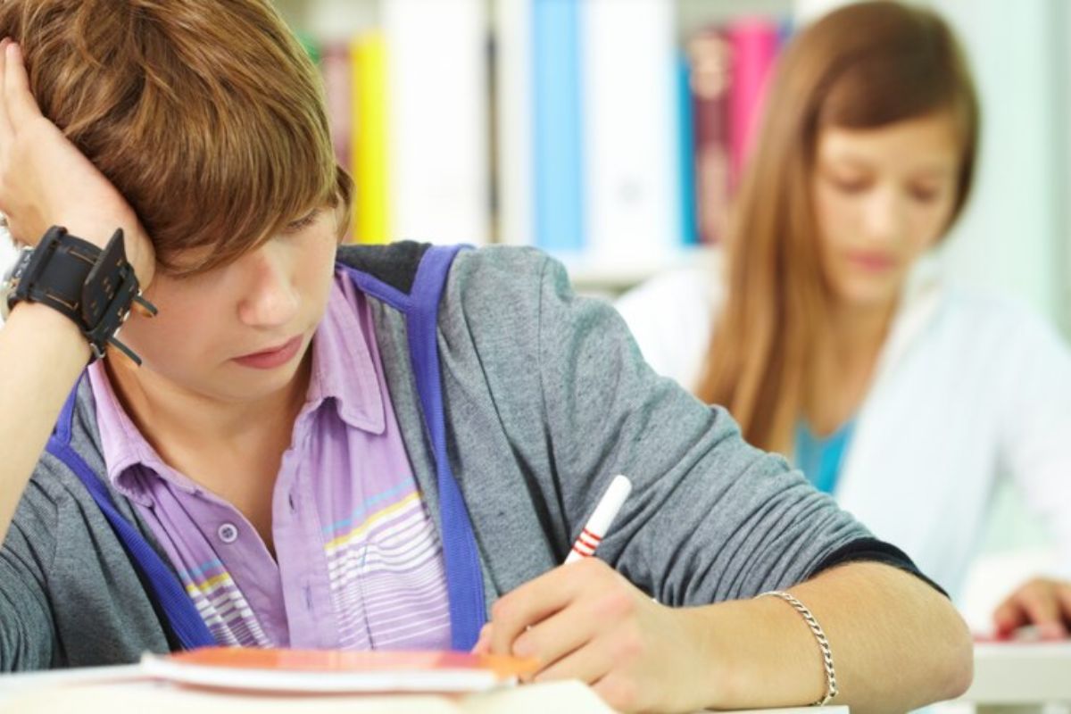 Um estudante com o cotovelo na mesa escrevendo no caderno. Ao fundo é possível ver outra aluna estudando
