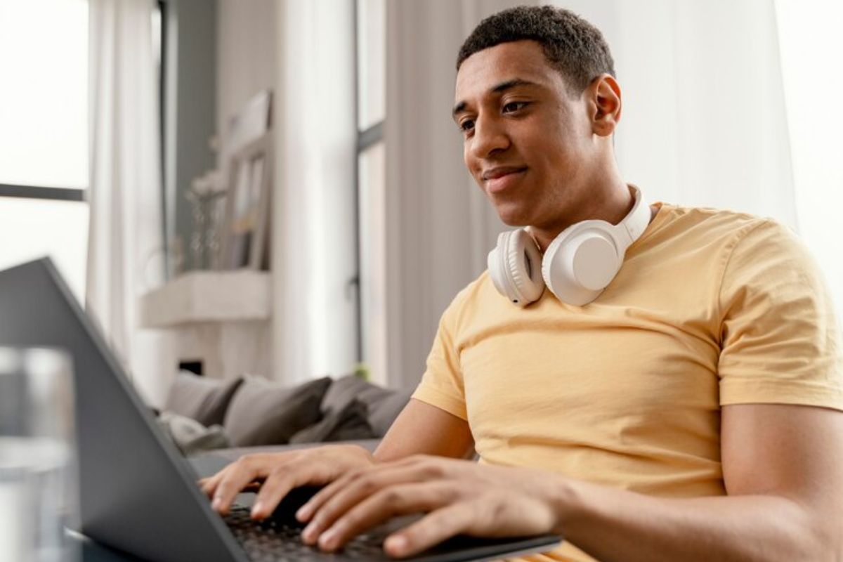 Um jovem dentro da sua casa com o um headphone no pescoço. Ele está sentado e mexe em sue notebook