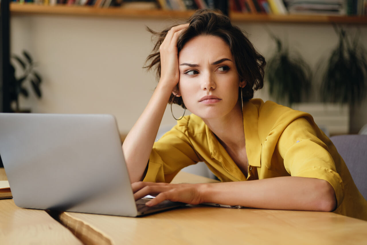 Jovem com expressão pensativa sentada diante o notebook com a mão na cabeça 