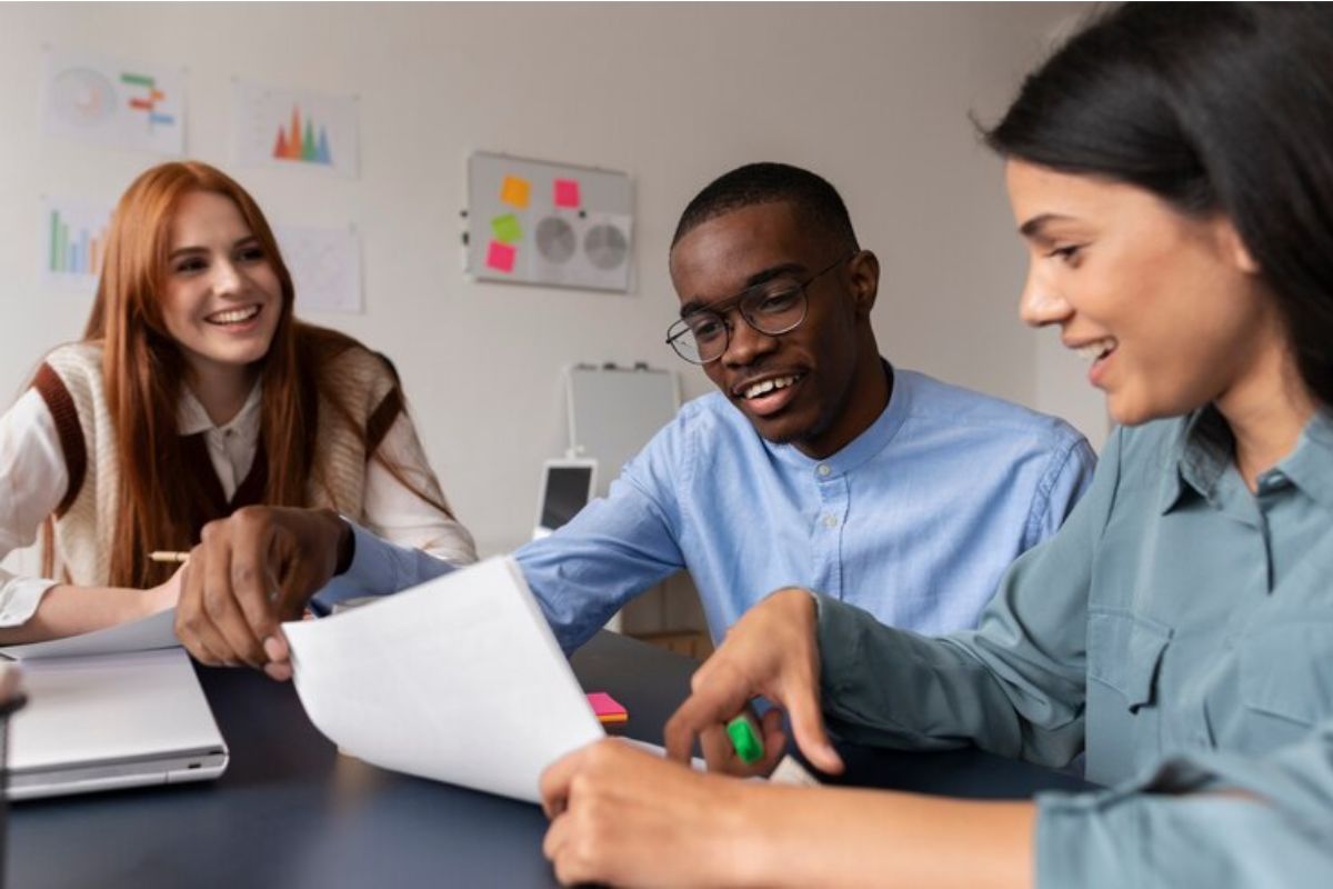 Um trio de estudantes em uma mesa. Eles estudam juntos no SENAC Itapetininga 2024. Uma estudante segura uma folha enquanto outros dois falam algo