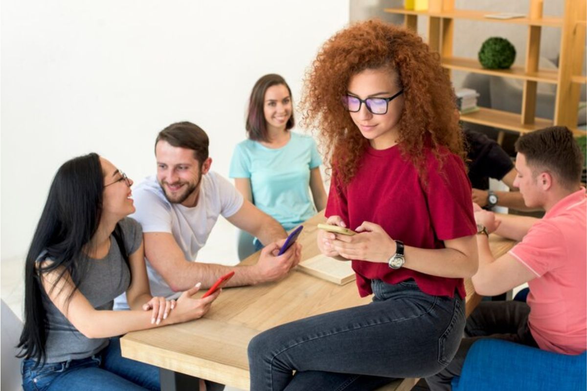 Um grupo de estudantes na sala de aula. Uma estudante está sentada em cima da carteira enquanto outros colegas estão em sua cadeiras enquanto conversam 