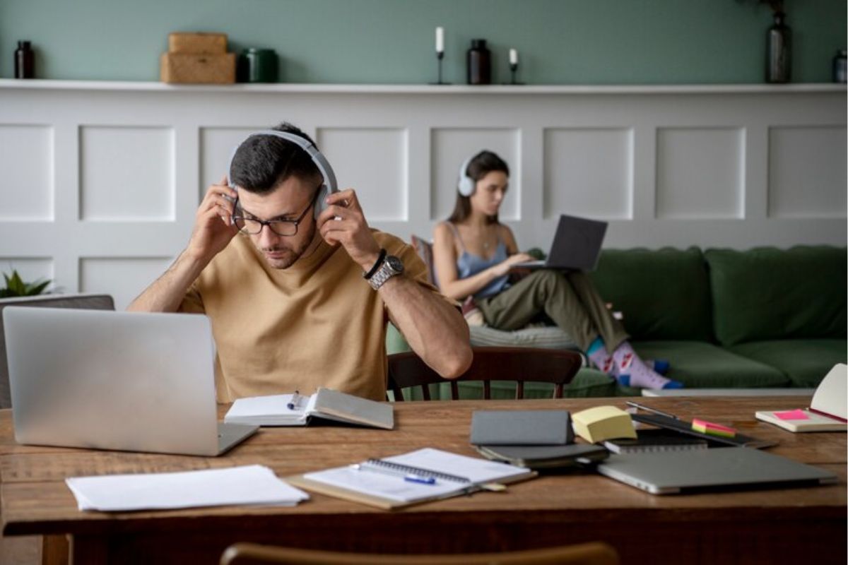 Dois alunos em casa. O homem está sentado em frente à mesa e sua namorada no sofá mais atrás. Ambos participam da Harvard Cursos Gratuitos 2024