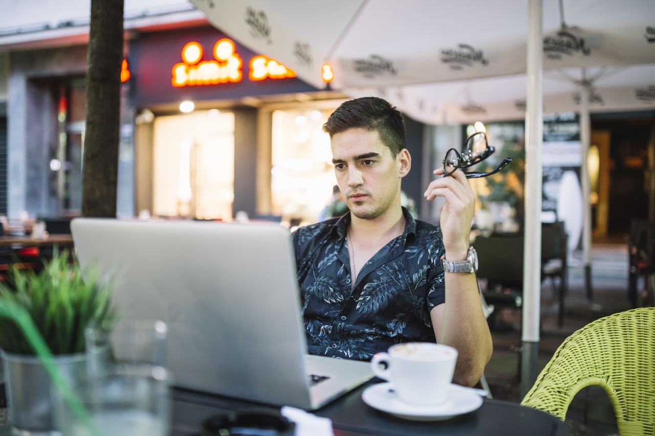 Jovem trabalha em seu notebook sentado na mesa de uma cafeteria 